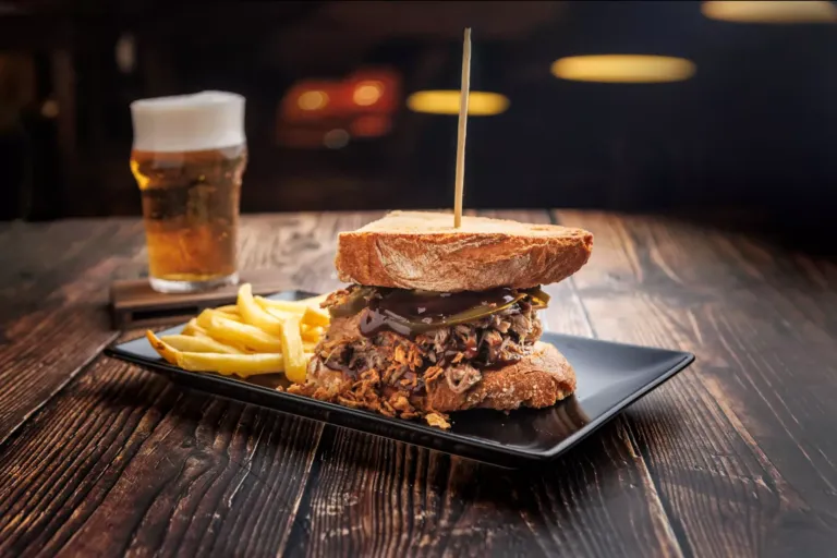 Sandwich de Pulled Pork Barbacoa, con carne de cerdo mechada, salsa barbacoa, jalapeños y cebolla crujiente; junto a una caña de cerveza