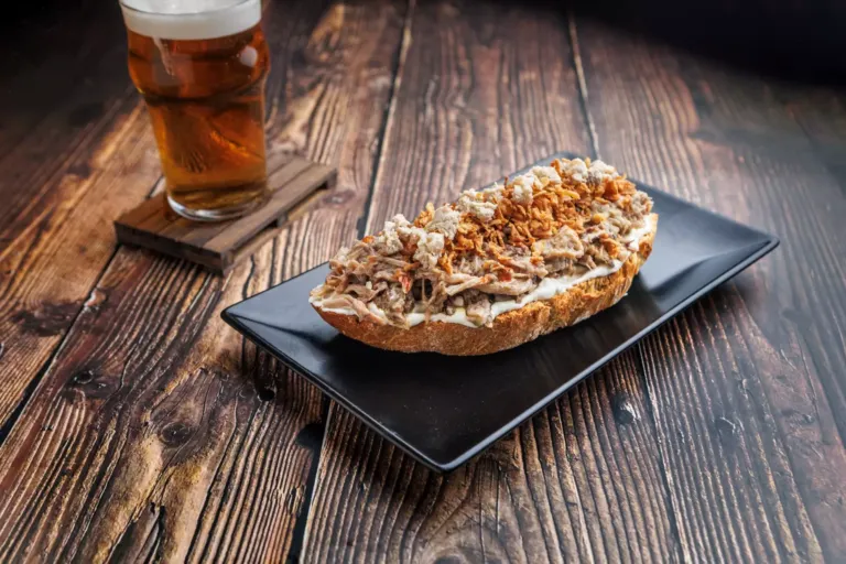 Caña de cerveza junto a un plato con una tosta de carne de cerdo mechada, alioli, queso azul, tabasco y cebolla crujiente