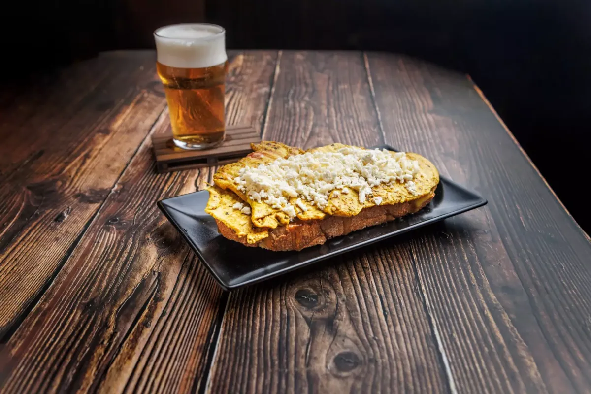 Tosta de pollo con pimienta y curry, queso de cabra y salsa de miel y mostaza junto a una cerveza